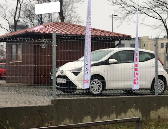 Auto Airport Samoobsługowy – Parking jest ogrodzony wysokim płotem. Biały samochód stoi przed drewnianym budynkiem. Obok powiewają flagi.