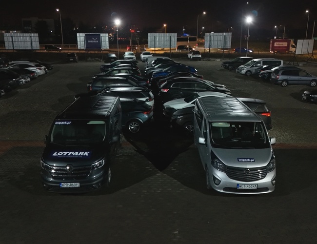 LotPark - Letiskové parkovisko. Povrch je osvetlený. Parkuje na ňom veľa osobných automobilov a dve autobusy.