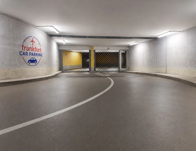 Podziemny parking z zaparkowanymi samochodami wzdłuż ściany, dobrze oświetlony przestronny korytarz.