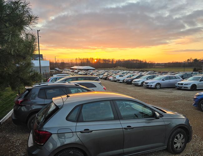 AleParking - Samochody stoją na żwirowym parkingu. W tle widać zachód słońca i drzewa.