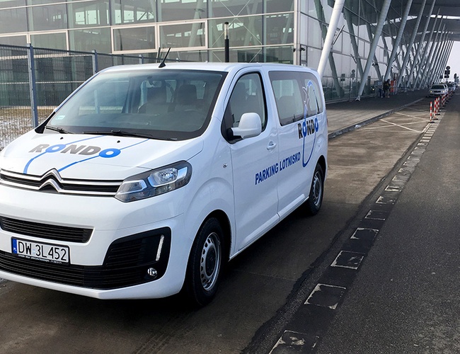 Parking Rondo – Biały van z logo obok lotniska. Auto stoi przy krawężniku. W tle widoczna szklana fasada budynku.