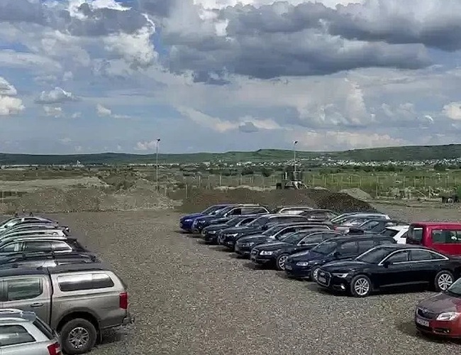 Szereg zaparkowanych samochodów na dużym żwirowym parkingu, otoczonym pagórkowatym terenem i niebem z chmurami.