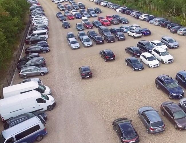 Balice Premium Park - Parking jest żwirowy i przestronny. Na placu zaparkowano wiele różnych aut. Teren otacza zieleń i ogrodzenie.