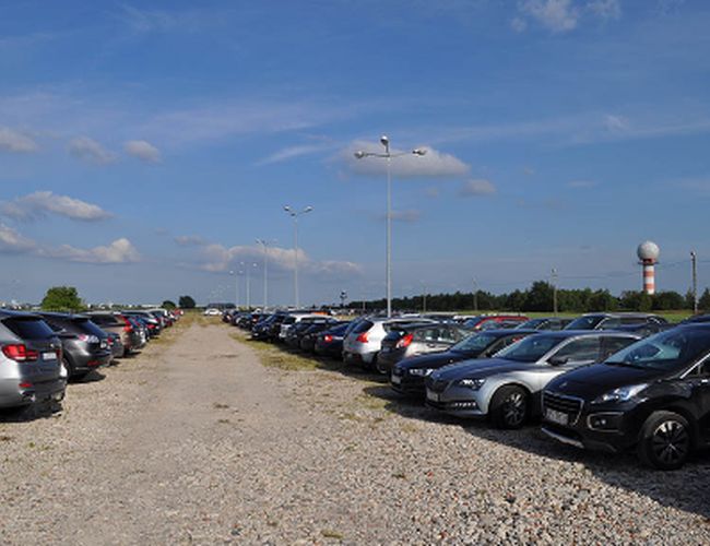 Parkplatz Flughafen – Auf dem Parkplatz stehen geparkte Autos. Das Gelände ist kieselig und beleuchtet. Im Hintergrund ist ein Radarturm zu sehen.