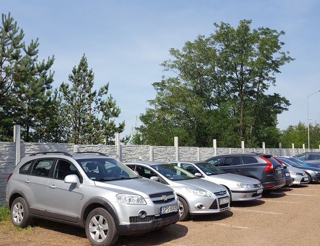 AAP-66 – Parking z ogrodzeniem i wieloma zaparkowanymi samochodami. W tle widać drzewa i latarnie. Na placu jest piaszczysta nawierzchnia.