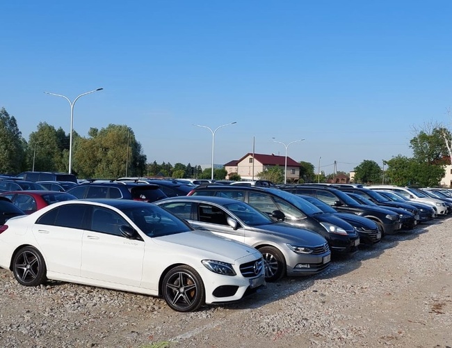 P12 – Gobierno de coches aparcados en superficie de grava. El aparcamiento es al aire libre con farolas. Al fondo se pueden ver casas y árboles.
