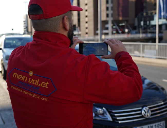 Parkovanie Mein Valet Vonku – Osoba v červenom kabáte robí fotografie auta. Na ulici stoja zaparkované autá. Na pozadí sa nachádzajú budovy.