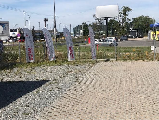 Auto Airport Samoobsługowy - Parking z kostką brukową i kamyczkami. Ogrodzenie z siatki, wokół materiałowe banery. W tle widać samochody.