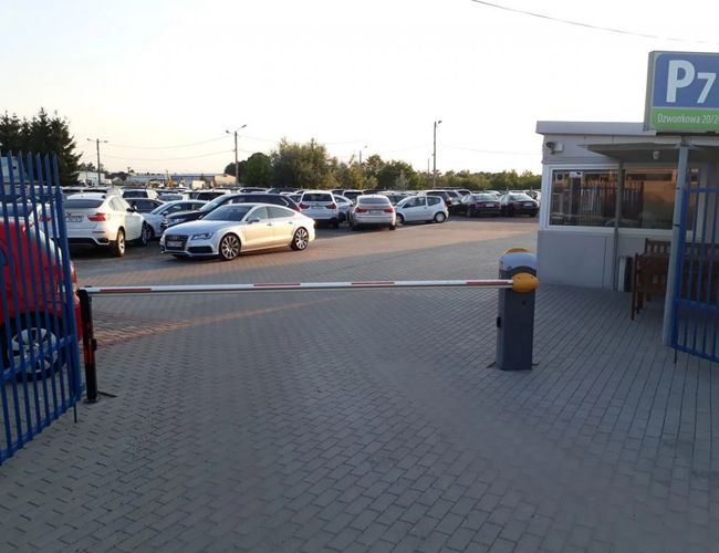 Parking P7 - Bramki otwierają się na przestronny plac. Widać wiele zaparkowanych samochodów. Teren jest otoczony ogrodzeniem.
