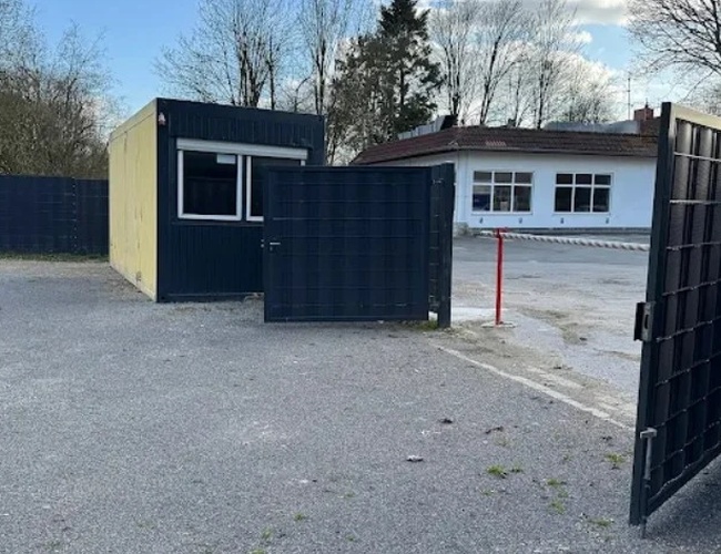 Parking Trust Park Fly – widoczny jest wjazd przez otwartą bramę. Po lewej stoi mały budynek. W tle znajdują się drzewa.