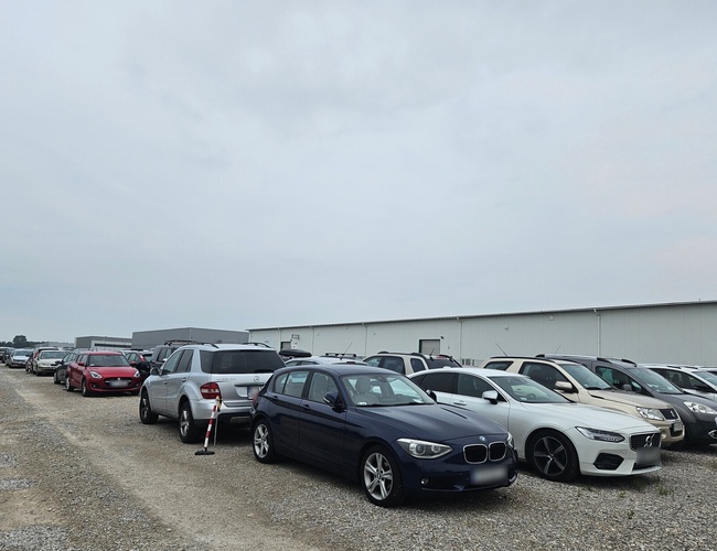 Holiday Parking – Na zdjęciu jest żwirowy plac parkingowy. Samochody stoją w rzędach. W tle widoczna jest hala.