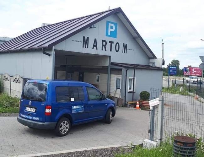 Parking MARTOM – Un coche azul está entrando en el área de estacionamiento. El edificio es de un solo piso y está cercado.