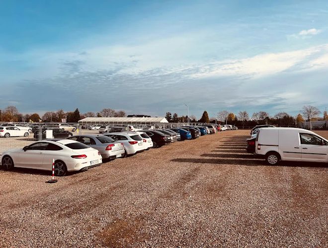 R8 Parking - Parking jest żwirowy i nieogrodzony. Znajduje się na nim wiele zaparkowanych samochodów. W tle widoczne są drzewa i budynki.