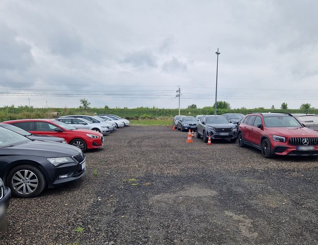 Parking Prime - Samochody ustawione na żwirowym placu. Obok stoją słupki drogowe. W tle widać zieleń i zarośla.