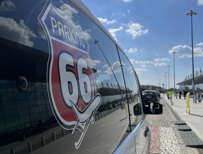 Estacionamiento 66 - Logo en la ventana del vehículo en el aeropuerto. De fondo se ve la terminal y el cielo con nubes.