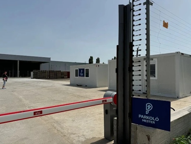 Szlaban wjazdowy na teren parkingu widziany z bliska, obok znajduje się ogrodzenie oraz biały kontener biurowy ustawiony na utwardzonym placu