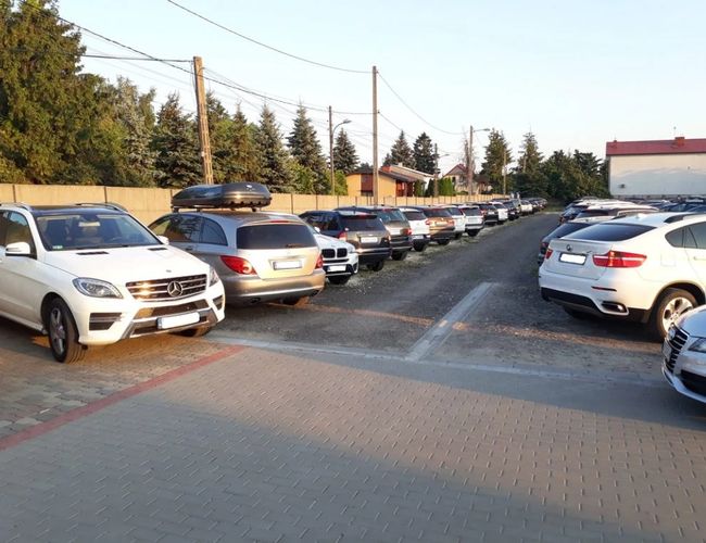 Parking P7 - Na parkingu stoją zaparkowane samochody. Teren jest utwardzony i otoczony drzewami. Widać ogrodzenie z tyłu.