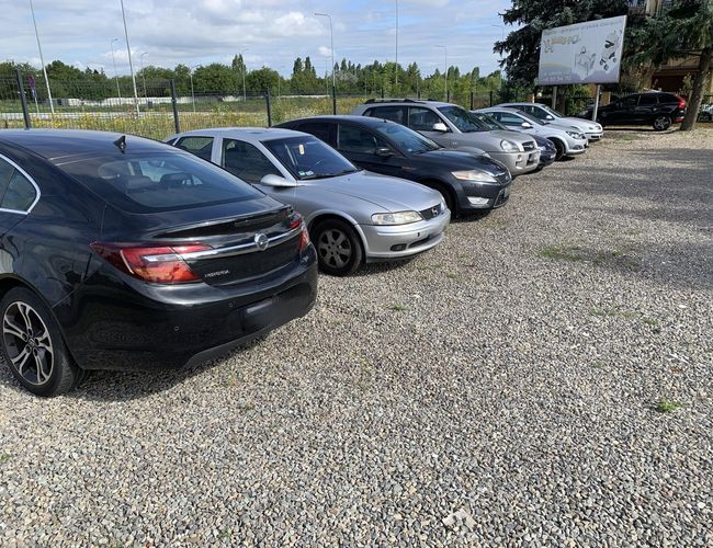 A1 Parking Max - Rząd samochodów stoi na żwirowanym placu. Parking jest ogrodzony siatką. W tle widać billboard.