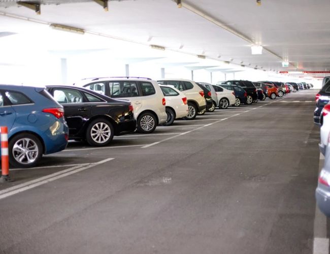 Wnętrze wielopoziomowego parkingu z samochodami zaparkowanymi po obu stronach drogi przejazdowej. Widoczne są oznakowane miejsca postojowe i oświetlenie sufitowe.