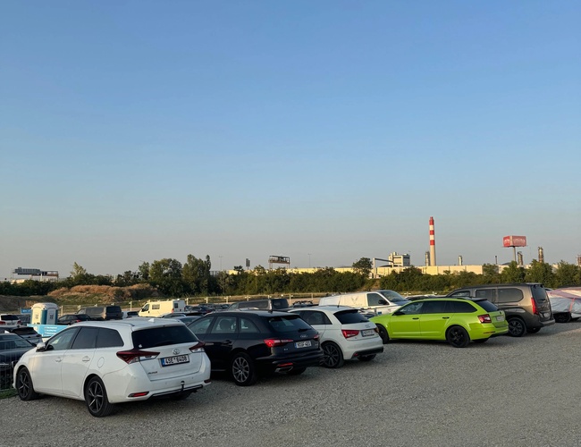Fila de coches aparcados en el estacionamiento. Superficie pavimentada visible y árboles al fondo.