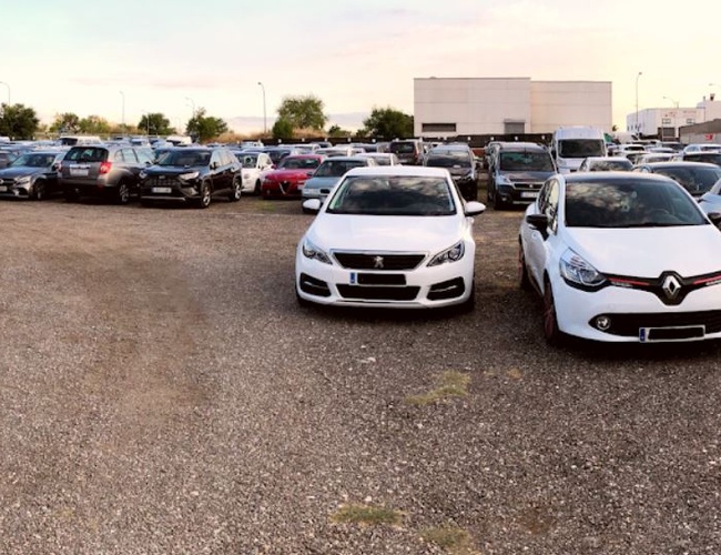 Parking Elite Valet - En el estacionamiento hay coches en una zona de grava. El área es abierta y no está cercada. En el fondo se puede ver un edificio.