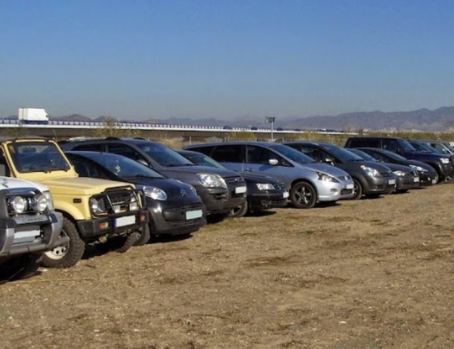 Parking PedroCar - Na ziemnym placu stoi rząd zaparkowanych samochodów. Nie widać ogrodzenia ani oświetlenia. W tle są góry.
