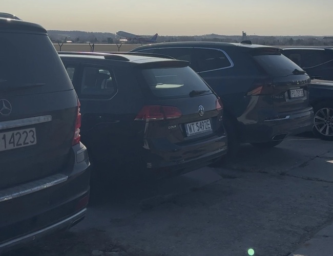 Parking MB Park – Samochody stoją na otwartym placu. W tle startuje samolot. Teren jest nieutwardzony.