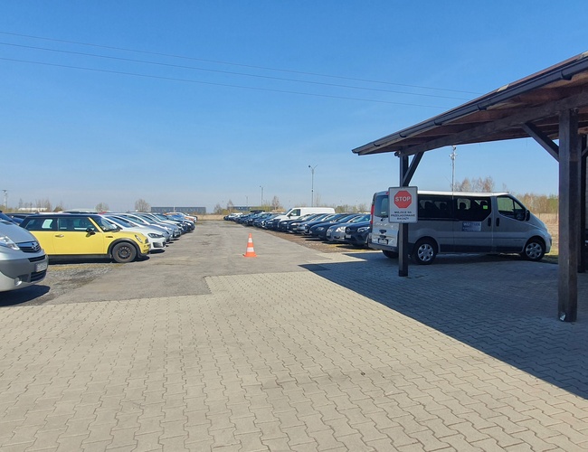 WIZ-PARK - Gran aparcamiento sin cercar de adoquines. Los coches están en filas. A la entrada hay un cobertizo techado.