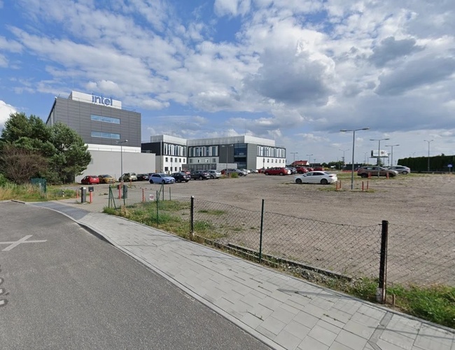 Parkovisko Piątka - Nezastavané parkovisko s sieťou, blízko kancelárskej budovy. Ulicou s chodníkom vedie k vchodu. V diaľke sú vidieť autá pod budovou.