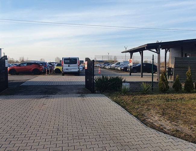 WIZ-PARK - El aparcamiento está cercado. La puerta está abierta. Se pueden ver coches aparcados y el edificio.