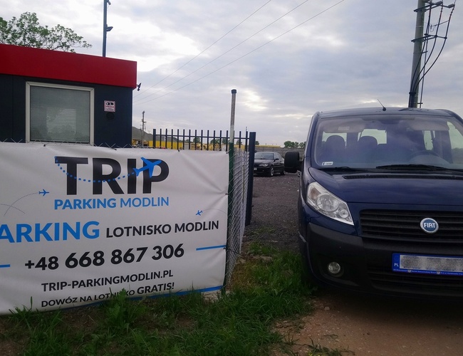 TRIP Parkovisko – Oplotené miesto s modrým autom pri bráne. Vedľa stojí šedý stánok s červenou strechou.