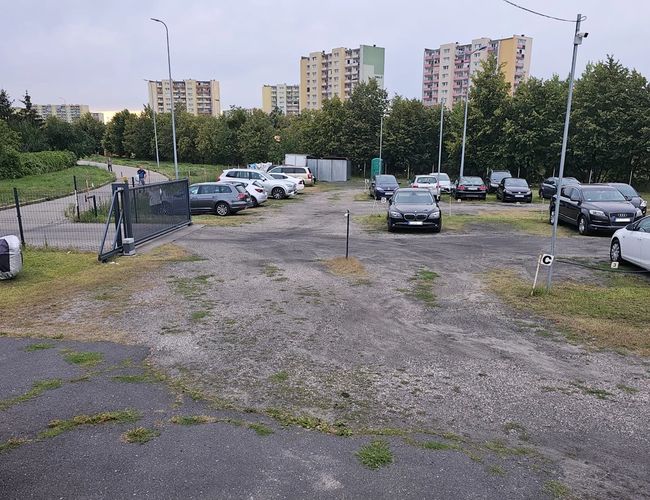 Estacionamiento BZG – El estacionamiento está rodeado de una cerca de metal. En el área hay coches aparcados. Al fondo se pueden ver edificios de apartamentos.