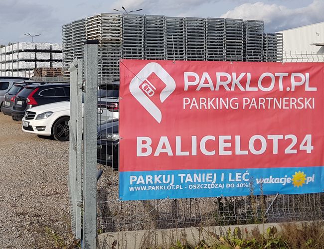 BaliceLot24 - Parking jest ogrodzony. W tle stoją zaparkowane samochody i stosy palet.