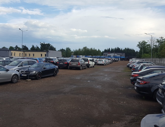 AAP-66 - Parking pełen samochodów, otoczony ogrodzeniem. W tle budynki i drzewa.