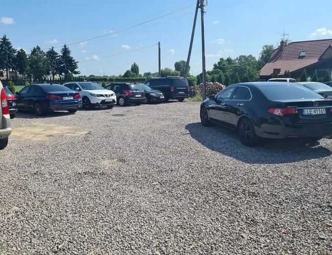 Parking Dominik - Na żwirowej nawierzchni stoi kilka samochodów. Otaczają go drzewa i domy. Jest słoneczny dzień.