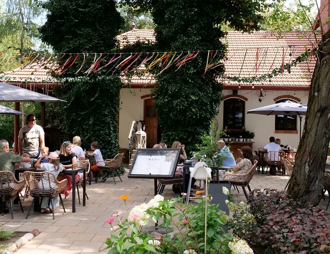 Parkovisko Zelený komplex Dwór Ostoya – Ľudia sedia v reštauračnej záhradke. Okolie lemujú rastliny a slnečníky.