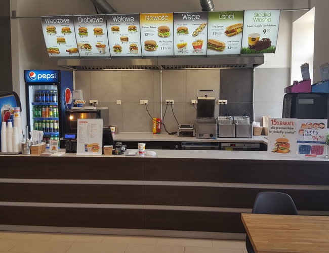 AAP-66 – Widoczna jest lada fast food z menu nad nią. Na ladzie znajdują się dystrybutory napojów oraz kubki. W tle widać sprzęt kuchenny.