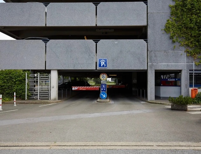 Wjazd do wielopoziomowego parkingu z zadaszonym wjazdem. Nad wjazdem znajduje się betonowa konstrukcja budynku, a po bokach widoczne są elementy zieleni i oznakowanie parkingu.