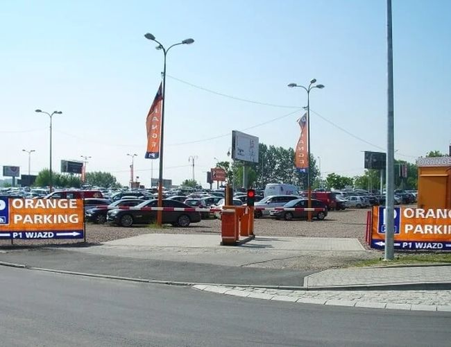 Orange Parking P3 - Parking jest na zewnątrz i dobrze oświetlony. Teren jest ogrodzony i widać liczne samochody. Na wjeździe są szlabany i znaki.