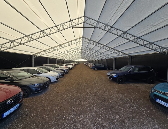Zakrytý parking AleParking - Mnohé autá pod strechou. Parkovisko je kamienité a ohradené. Konštrukcia strechy z kovových nosníkov.