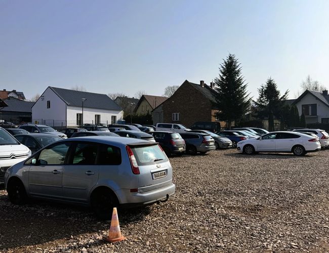 Parking Amber - Na żwirowym terenie stoją zaparkowane samochody. W tle widać budynki i drzewa.