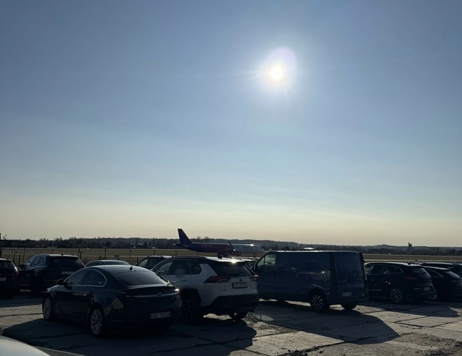 Parking MB Park - Kilka samochodów stoi na betonowej nawierzchni. Na horyzoncie widać samolot. Słońce świeci na jasnym niebie.