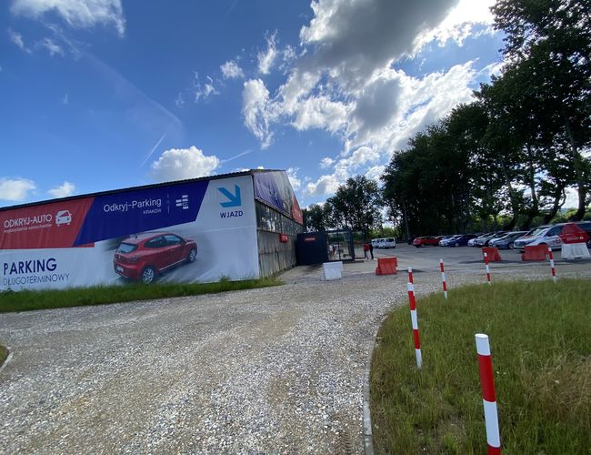 Odkryj-Parking - Żwirowa droga prowadzi do ogrodzonego parkingu. W tle stoją zaparkowane samochody. Drewniane barierki oddzielają teren.