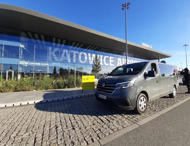 GoPark – Frente a la terminal B del Aeropuerto de Katowice hay un autobús gris. La superficie pavimentada lleva a la entrada.