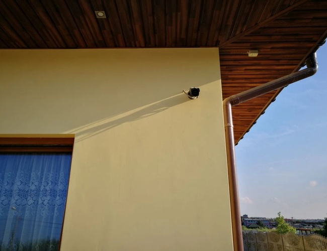 Parking Darex - Pared blanca del edificio con cámara. Tubería visible y fragmento de ventana. Tejado de madera y cielo al fondo.