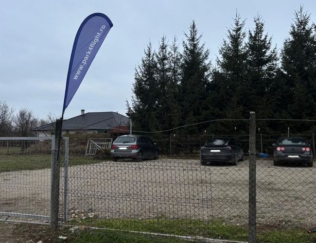 Wjazd na parking Park4flight Maramureș z otwartą metalową bramą, flagą informacyjną przy ogrodzeniu oraz kilkoma samochodami zaparkowanymi na przestronnym, utwardzonym placu w pobliżu lotniska.