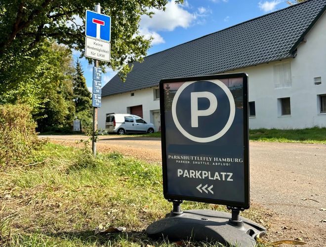 ParkShuttleFly Hamburg - w centrum stoi znak parkingu, w tle samochód stoi na żwirowym placu. W tle widoczne są drzewa i budynek.