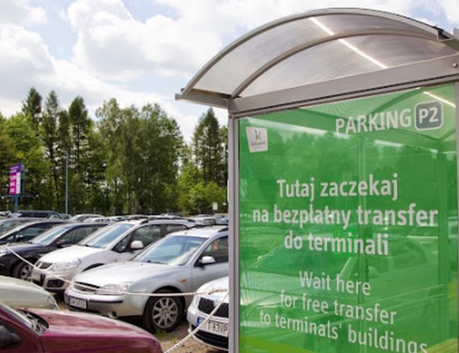 Oficjalny Parking P2 - Rząd zaparkowanych samochodów. Przystanek z informacją o transferze do terminali. W tle drzewa i niebo.