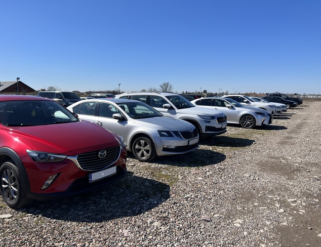 Aparcamiento P12 - En el terreno de grava hay una fila de coches aparcados. El área está abierta y al fondo se ve un edificio.