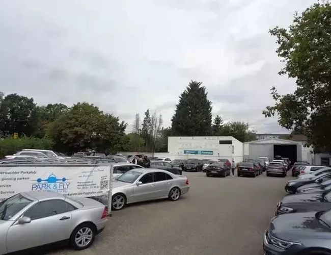 Park&Fly Düsseldorf Parkbereich - Der Bereich ist voll geparkter Autos. Umgeben von Bäumen, im Hintergrund ist ein Gebäude zu sehen.